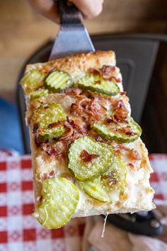 a person holding a slice of pizza with cucumber slices on it and a knife in their hand