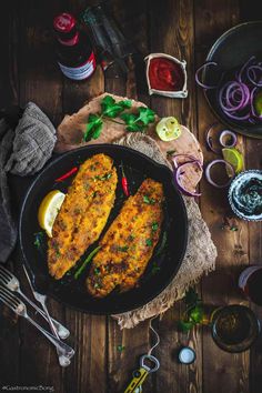 two fish fillets in a skillet with vegetables and sauces on the side