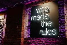 a neon sign that says who made the rules? on a brick wall with purple lighting