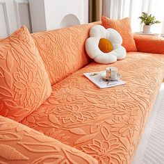 an orange couch with decorative pillows and a coffee cup on it in front of a window