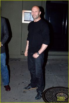 bald man in black shirt and jeans standing on the sidewalk with his hands in his pockets