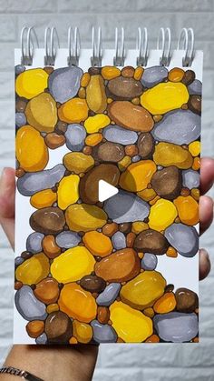 a hand holding up a spiral notebook with rocks on the front and side, which are painted in different colors