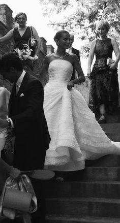 a woman in a wedding dress walking down some steps with other people around her and one man holding a purse