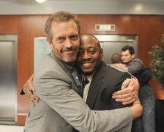 two men hugging each other in an office setting with people standing around the room behind them