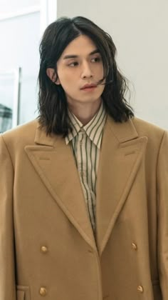 a man with long black hair wearing a tan coat and striped shirt standing in front of a white wall