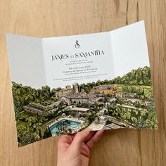 a person holding up a wedding program in front of a wooden table with trees and buildings