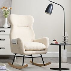 a white rocking chair next to a black table with flowers on it and a lamp in the corner