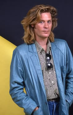 a man with long hair standing in front of a yellow and black background wearing a blue jacket