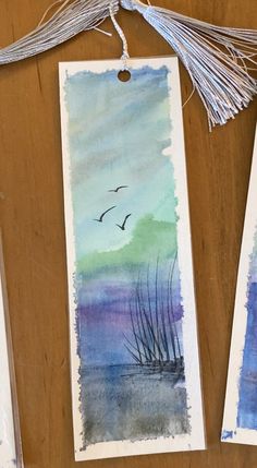 two bookmarks with watercolors on them hanging from a wooden table next to some tassels