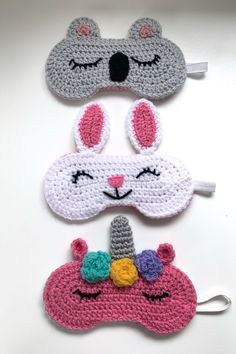 three crocheted sleeping masks with bunny ears and an unicorn face on them, one is