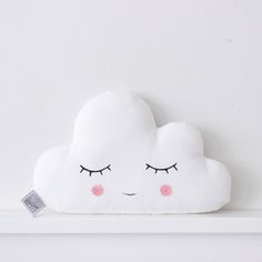 a white cloud pillow with pink cheeks and eyelashes on it's face sitting on a shelf