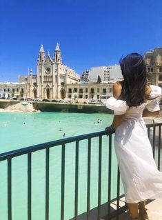 a woman in white dress looking out over the water from a balcony overlooking buildings and beach