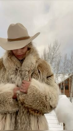 a woman wearing a fur coat and hat standing in the snow with her arms crossed