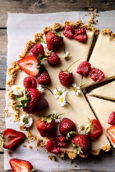 a cheesecake topped with strawberries and flowers on top of a wooden table next to a piece of pie