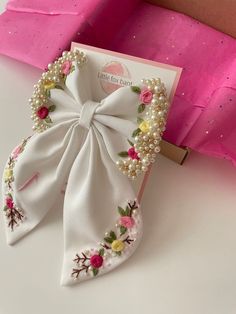 a white bow with flowers and pearls on it sitting next to a pink gift bag