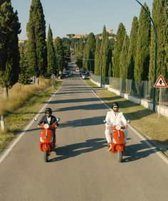 two men riding on the back of red scooters down a long country road