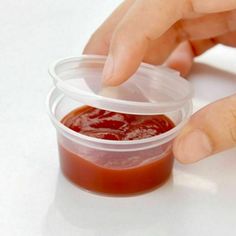 a hand reaching for a plastic container with ketchup in it on a white surface