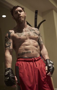 a shirtless man with tattoos and boxing gloves standing in front of a mirror wearing red shorts