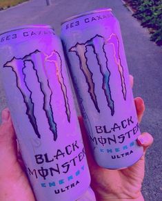 two cans of monster energy drink in the palm of someone's hand, both have purple and black lightning prints on them