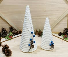 two white knitted christmas trees with blue beads and pine cones on the table next to each other