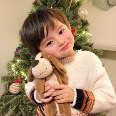 a young boy holding a stuffed dog in front of a christmas tree