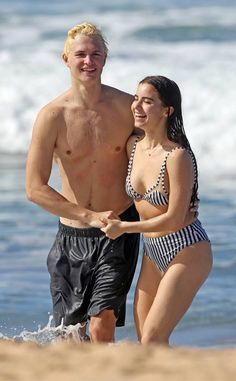 a man and woman in swimsuits walking on the beach with waves behind them