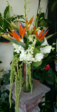 a vase filled with white and orange flowers on top of a stone pedestal next to plants