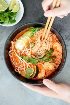 someone holding chopsticks over a bowl of noodles and vegetables with shrimp, cucumber, and lime