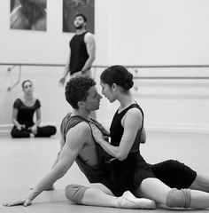 a man and woman sitting on the floor in a dance studio with dancers behind them