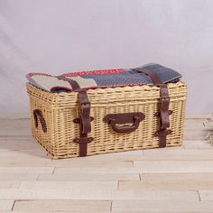 a wicker picnic basket with leather straps on the floor next to a potted plant