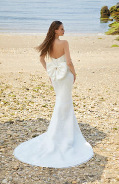 a woman is standing on the beach in a white dress with a bow at her back