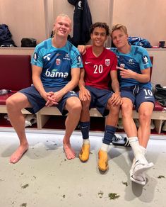 three soccer players sitting on a bench in the dressing room with their arms around each other