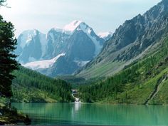 the mountains are covered with snow and green trees in front of a body of water