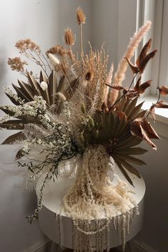 an arrangement of dried flowers and plants on a white table in front of a window