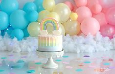 a birthday cake with a rainbow on top and balloons in the back ground behind it