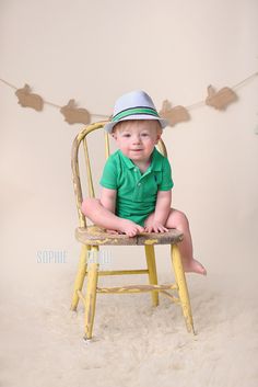 a baby sitting in a yellow chair wearing a green shirt and hat with leaves on the wall behind him