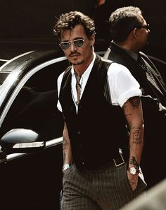 a man with tattoos and glasses standing in front of a car wearing a vest, tie and sunglasses