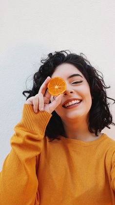 a woman holding an orange slice up to her face and smiling with one hand on her head