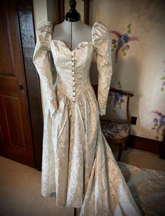 This renaissance-style dress is truly unique. The fitted princess line bodice has a square neckline front and back with a small vee at center front. This is accentuated by a mock opening in the front with "pearl" buttons, each encircled with rhinestones. Some of the buttons are missing a rhinestone. Puff sleeves, with elastic at the shoulders for wearing ease, taper to points at the wrist with a four button and loop closure. The drop waist come to a point in the front and back. Skirt is gathered. Zipper closure. What makes this truly renaissance looking is the fabric. It has gold metallic slightly raised flowers on a cream background. Dress has been cleaned and preserved since the 1991s and is in excellent condition.  Not only would this make a wonderful wedding dress, it would also be ama Elegant Gown With Fitted Bodice And Heart-shaped Neckline, Heart-shaped Neckline Gown With Fitted Bodice For Debutante Ball, Elegant Ball Gown With Lined And Fitted Bodice, Fitted Gown With Heart-shaped Neckline For Debutante Ball, Fitted Gown With Boned Bodice And Sweetheart Neckline, Fitted Bodice Ball Gown With Corset Back, Corset Dress With Empire Waist And Lined Bodice, Fitted Empire Waist Corset Dress With Lined Bodice, Formal Gown With Fitted Bodice And Sweetheart Neckline