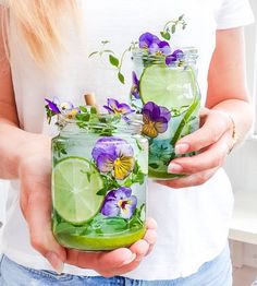 two women holding mason jars with flowers and limes in them, one is filled with water