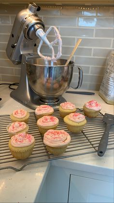 some cupcakes are sitting on a rack in front of a mixer with pink frosting