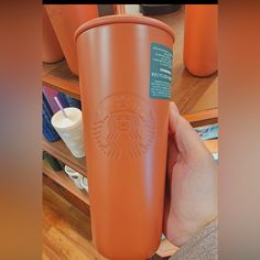 a hand holding an orange starbucks cup in front of some cups on a wooden shelf