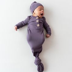 a baby laying on its back wearing a purple outfit with buttons and a beanie