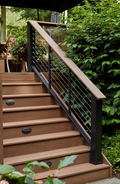 a wooden deck with metal railing and handrails in front of some trees, bushes and flowers