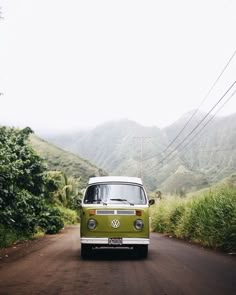 an old vw bus is driving down the road in front of some mountains and trees