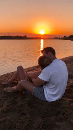 a man and woman are sitting on the sand by the water as the sun sets