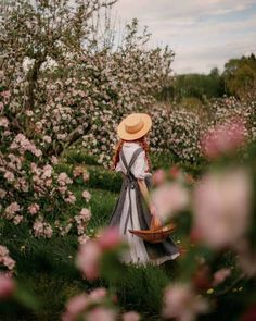 a woman wearing a hat and carrying a basket walks through an orchard full of pink flowers