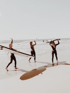 three women in wetsuits carrying surfboards on the beach with their arms up