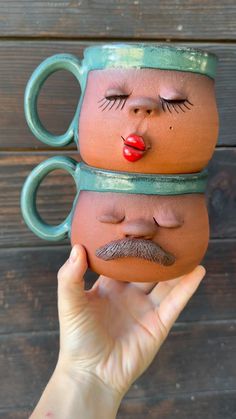 a person holding two coffee mugs with faces painted on them, one has a mustache and the other has eyes closed