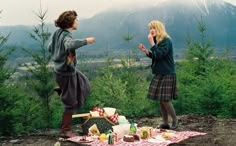 two women standing in front of a picnic table with food and drinks on the ground